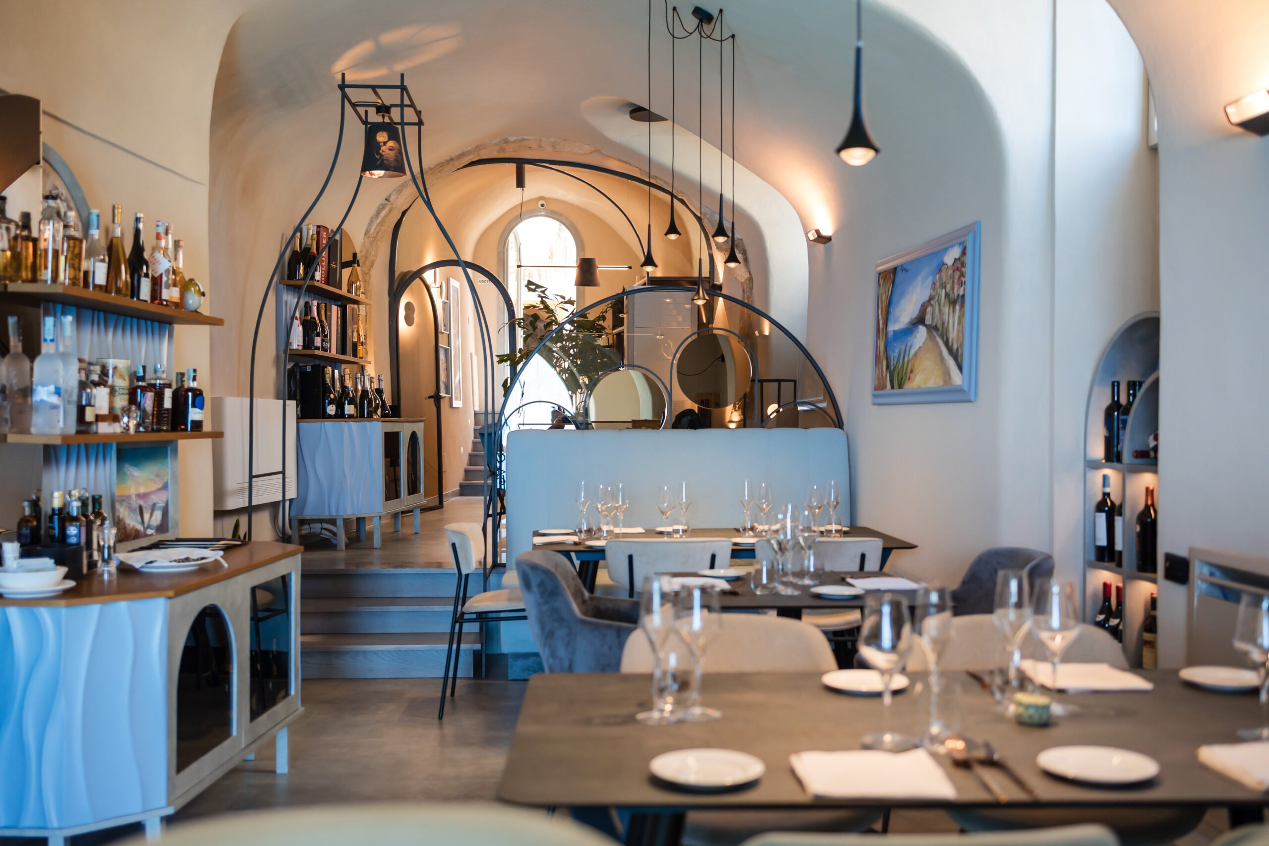 Gli interni del ristorante Deodato Lounge Bistrot di Tropea, sulla destra alcuni tavoli apparecchiati in stile minimal ed elegani, sulla sinistra due mobiletti di design con sopra bottiglie di cgampagne