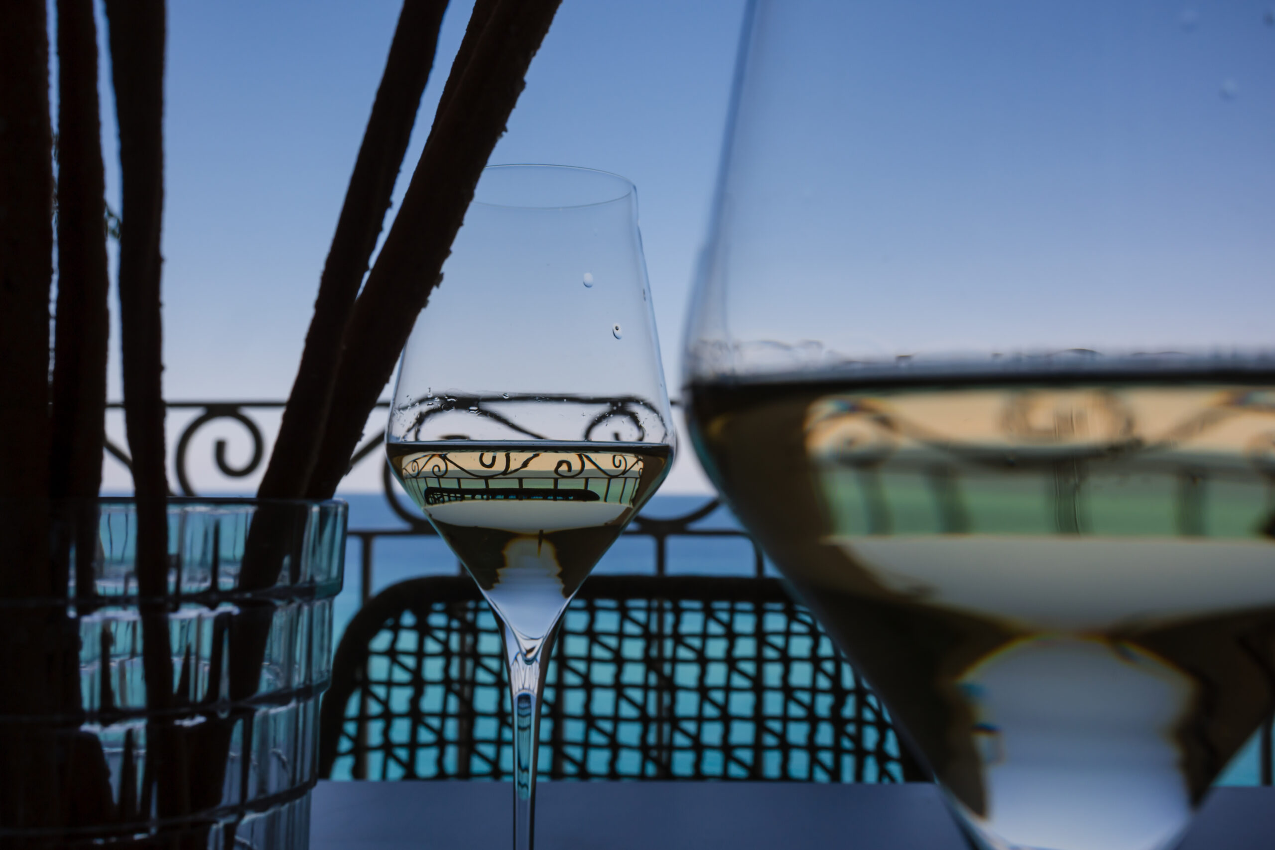 un primissimo piano di due calici di vino bianco freschi su un balcone affacciato dalla rupe di Tropea. I calici sono poggiati su un tavolino con due sedie all'ora del tramonto