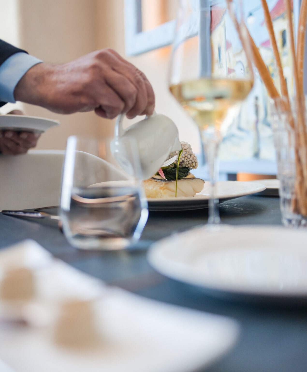 tavola imbandita con un calice di vino bianco, un bicchiere di acqua e una mano che versa una salsa su un piatto di Baccalà gourmet